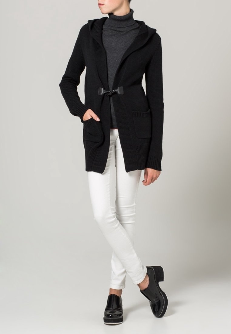 Czarny dzianinowy kardigan z kapturem, z zapięciem na sznurki i dwoma przednimi kieszeniami, zestawiony z białymi dopasowanymi spodniami i czarnymi butami.