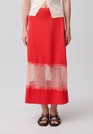 Donna che indossa una gonna midi rossa con un pannello in pizzo trasparente sopra l'orlo e sandali neri con cinturini, in piedi davanti a uno sfondo semplice.