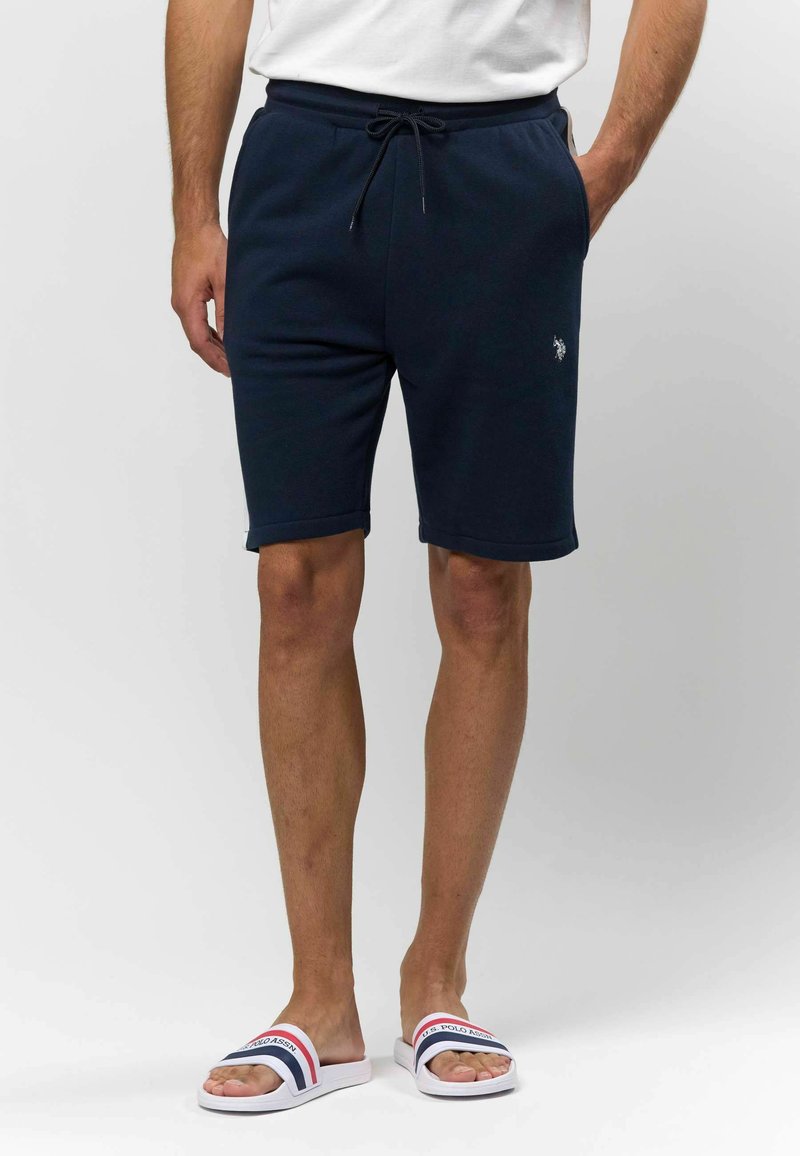 Shorts en coton bleu marine avec une taille élastique, un cordon de serrage et des poches latérales. Présente un petit logo sur la jambe gauche. Associés à des sandales blanches à glissière.