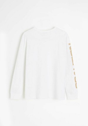 Biała koszula z długim rękawem, widziana od tyłu, z beżowymi blokowymi literami biegnącymi pionowo wzdłuż prawego rękawa.