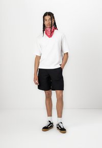 T-shirt blanc à manches courtes associé à un short noir, complété par un bandana rouge autour du cou et des baskets noires avec une semelle en caoutchouc.