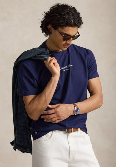 Άνδρας με γυαλιά ηλίου, μπλε σκούρο t-shirt Ralph Lauren, λευκό παντελόνι με καφέ ζώνη, κρατώντας ένα τζιν μπουφάν στον ώμο του.