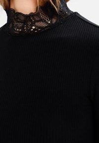 Nærbillede af en person iført en sort ribbet top med en høj, intrikat sort blondekrave og rødt hår synligt ovenfor.