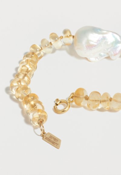 Βραχιόλι με διάφανα κεχριμπαρένια χάντρες, ακανόνιστη μεγάλη λευκή μαργαριταρένια χάντρα, χρυσό κούμπωμα και μικρή χρυσή ταμπέλα με χαραγμένο το όνομα "Loern Stewart".