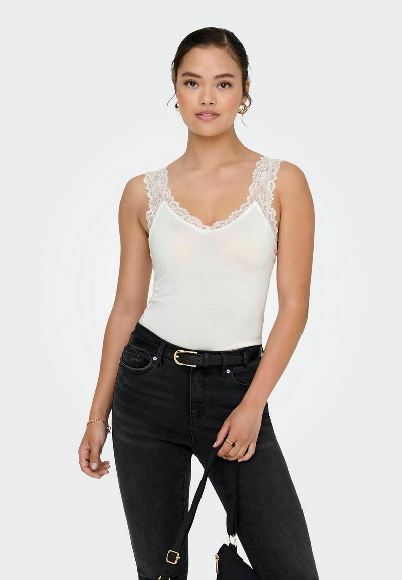Hvid tanktop med blondedetalje, v-hals og tætsiddende design. Sammen med højhalset sorte jeans og en sort taske. Minimalistisk stil.