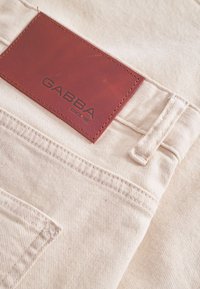 Beige denim fabric featuring a brown leather patch with "GABBA" embossed and stitching details, showcasing a textured surface and tailored design.