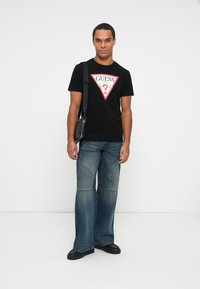Tricou negru cu un logo grafic triangular roșu, asociat cu blugi largi de culoare albastră deschisă și pantofi negri. Genți mici de umăr negre.