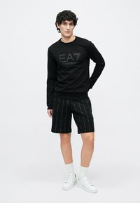 Νεαρός άνδρας στέκεται με το ένα χέρι πίσω από την πλάτη, φορώντας μαύρο φούτερ EA7, μαύρα σορτσάκια με σχέδια, λευκές κάλτσες και λευκά αθλητικά παπούτσια.