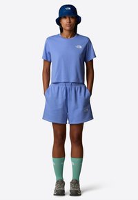 Maglietta cropped in cotone azzurro chiaro e pantaloni corti abbinati, con tasche laterali e logo. Abbinata a calzini verdi e un cappello a secchiello blu.