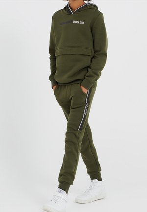 Dítě má na sobě olivově zelenou mikinu s kapucí a odpovídající tepláky s bílými pruhy, doplněné bílými kotníkovými teniskami, stojí s rukama v kapsách.