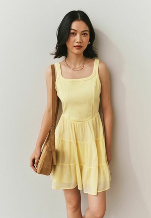 Fiatal nő sárga ujjatlan ruhában, réteges szoknyával, barna válltáskát viselve, egy sima világos fal előtt áll.