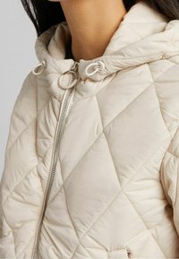 Lehká prošívaná bunda v béžové barvě. Obsahuje kapuci se stahovacími šňůrkami, plný přední zip a prošívání v diamantovém vzoru pro texturu.