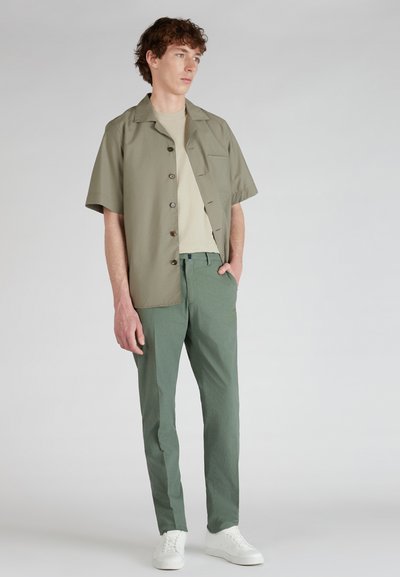 Camicia a maniche corte color oliva con bottoni marroni, t-shirt beige sotto, pantaloni verdi strutturati e sneakers bianche. Design minimalista.