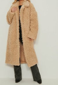 Beige keinoturkistakki, jossa on pörröinen pinta, pitkät hihat, avoin etuosa ja rento istuvuus, yhdistetty tummiin farkkuihin ja kärjestä käyrät mustat kengät.