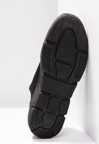 Semelle de basket noire avec des motifs en caoutchouc texturé reposant contre une surface d'angle blanche.