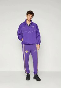 Ung mand iført et lilla Los Angeles Lakers træningssæt med sorte sneakers, stående mod en ensfarvet lys baggrund.