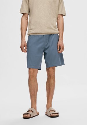 Light beige relaxed-fit short-sleeve shirt, paired with blue cotton shorts and beige slip-on sandals. Casual, comfortable outfit.