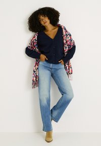 Cárdigan azul marino con botones sobre un envoltorio floral colorido, combinado con vaqueros azul claro y botas cortas color canela. Textura suave, ajuste cómodo.