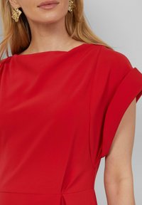 Robe rouge avec une texture lisse, des manches courtes retroussées et une coupe ample. Les détails notables incluent un subtil twist à la taille.