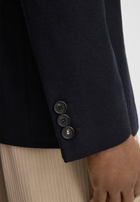 Granatowy wełniany rękaw marynarki z trzema czarnymi guzikami z błyszczącym akcentem. Tkanina ma gładką teksturę, podkreślającą dopasowany fason.