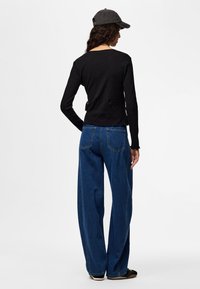 Kobieta w czarnej bluzce z długim rękawem, szerokich niebieskich dżinsach, czarnych butach i ciemnej czapce z daszkiem, stoi tyłem na jednolitym tle.