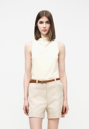 Jeune femme aux cheveux bruns raides portant un haut sans manches crème, un short beige retroussé et une ceinture marron, debout devant un fond blanc.