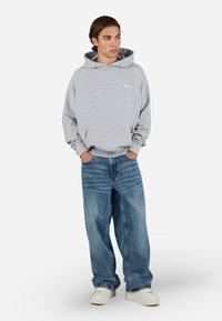 Grå hættejakke med snor, prydet med et logo foran, parret med løse blå jeans og hvide sneakers. Baggrunden er ensfarvet hvid.