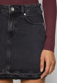 Jupe en denim noir à la silhouette ajustée, dotée de poches frontales, d'une fermeture à bouton métallique et d'un détail d'ourlet effiloché.