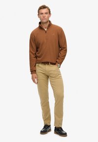 Brauner Quarter-Zip-Pullover, beiger Chino und dunkle Schuhe. Der Pullover hat eine strukturierte Oberfläche. Der Gürtel hat ein geflochtenes Muster.