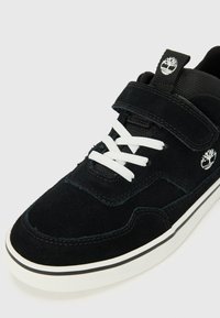 Baskets en suede noir avec lacets blancs, bande Velcro et logo. Comprend une semelle en caoutchouc blanc avec bord noir et un tissu texturé au niveau du col.
