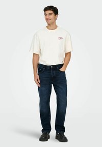 Camiseta de algodón color beige claro con texto rojo, combinada con jeans de mezclilla azul oscuro. Modelo vistiendo zapatos negros, de pie con las manos en los bolsillos.