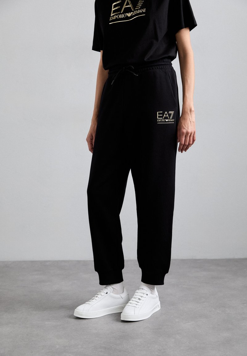 Zwarte sweatpants met elastische boorden en een trekkoord in de taille, met een gouden EA-logo. Gecombineerd met witte sneakers.