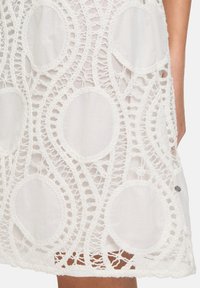 Weißes Spitzenkleid mit einem kreisförmigen und wellenförmigen Häkelmuster, einer halb durchsichtigen Textur und einem bestickten Akzent am Saum.