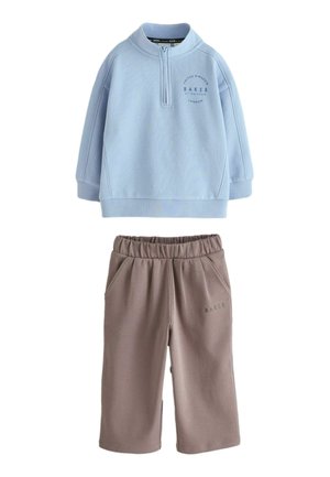 Hele sinine pika varrukaga pullover poole lukuga ja pruunid elastse vöökohaga laia säärega püksid, mõlemal väikeste tekstilogodega detailid.