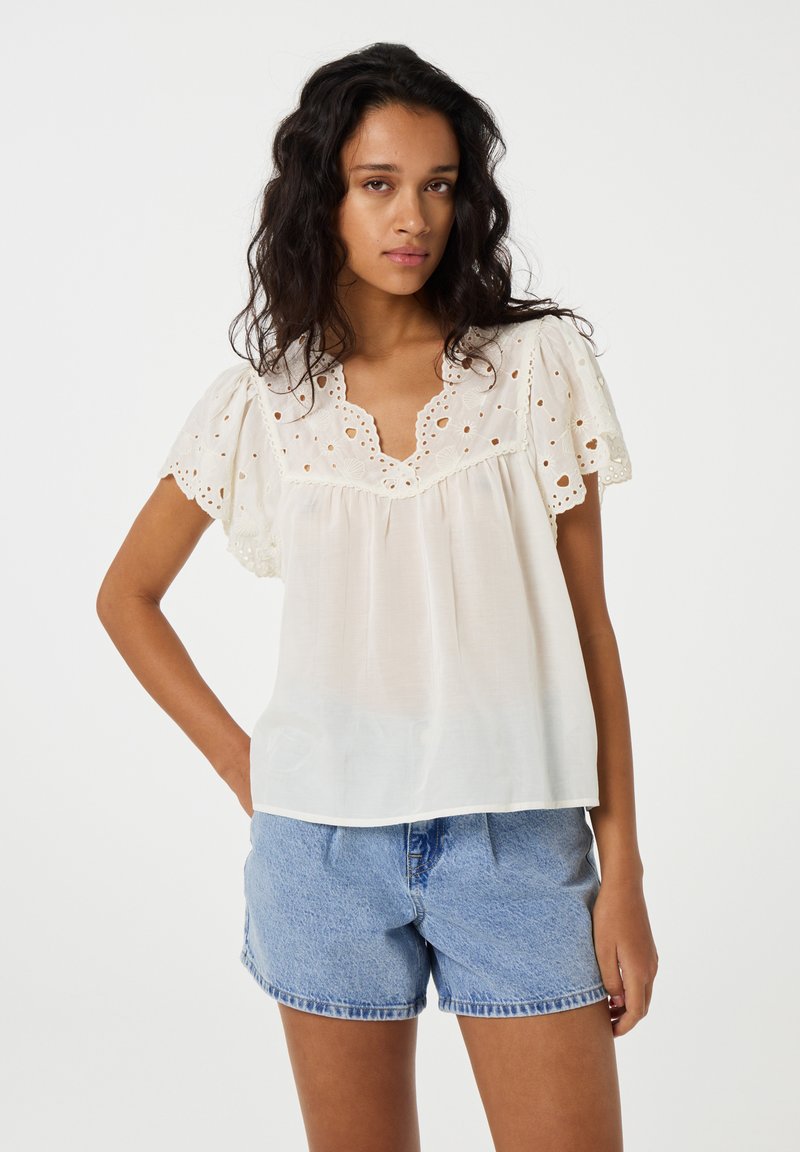 Witte blouse met borduursels en geschulpte mouwen, met een V-hals en een vloeiende pasvorm. Gecombineerd met lichtblauwe denim shorts.