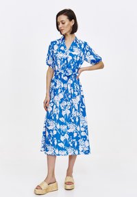 Niebieska sukienka midi w kwiatowy wzór z krótkimi rękawami, z detalem w postaci związania w talii oraz lekkiego materiału, zestawiona z plecionymi sandałami na platformie.