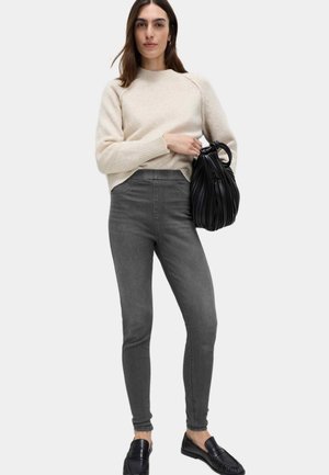 Žena v krémovém svetru, šedých úzkých džínách, černých mokasínách, držící černou plisovanou kabelku na jednoduchém světlém pozadí.