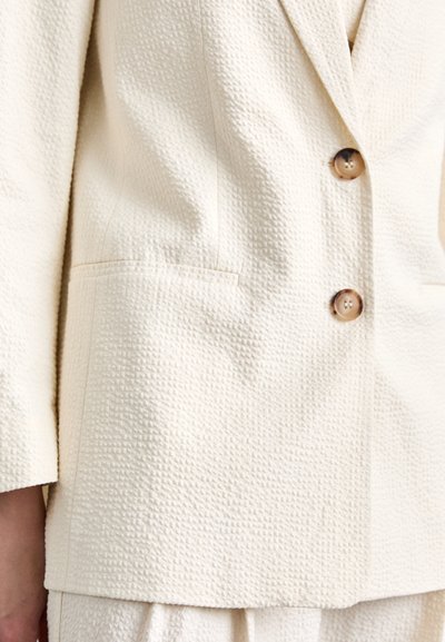 Crèmekleurige blazer in textiel met structuur, voorzien van een geknipt revers, twee knopen en een enkel zakje. Gladde knoopdetails zorgen voor een accent.