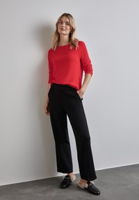 Maglione rosso a maniche lunghe, vestibilità comoda; pantaloni neri a gamba larga; mocassini neri con dettagli morbidi; sfondo neutro con pavimentazione grigia.