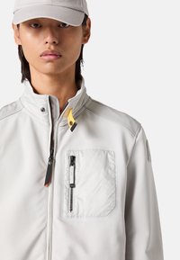 Hellgraue Jacke mit hohem Kragen, mit Reißverschlüssen versehenen Taschen und schwarzen Akzenten. Sie zeichnet sich durch eine glatte Textur und ein minimalistisches Design aus.