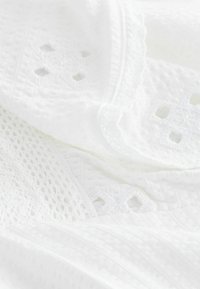 Tissu blanc avec des motifs complexes de dentelle et de broderie à œillets, montrant des textures douces et des dessins découpés délicats.