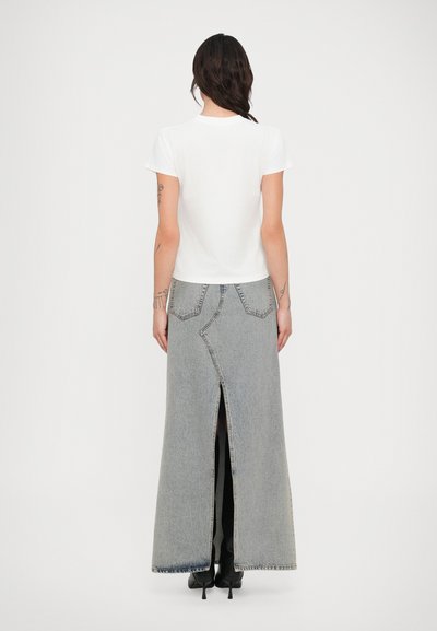 Nő hosszú sötét hajjal, fehér rövid ujjú pólót, világosszürke maxi farmer szoknyát hátul hasítékkal, és fekete magassarkú csizmát visel.