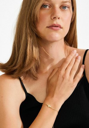 Kuldne käevõru, millel on peen ja keerukas muster. Modell kannab musta soonikutest topi. Juuksed on pikad ja lainelised, nägu ümbritsevad.