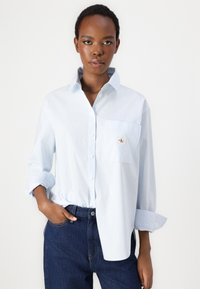 Light blue and white striped button-up shirt with a pocket and logo detail, made from a smooth fabric; long sleeves rolled at the cuffs.