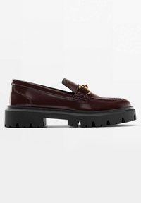 Mocassin en cuir marron foncé avec semelle épaisse à crampons noirs et boucle décorative dorée sur le dessus, vue de côté sur fond blanc.