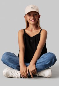 Top de tirantes finos negro texturizado, con un pequeño logo, combinado con jeans de color azul claro y zapatillas blancas, sentado sobre un fondo gris.
