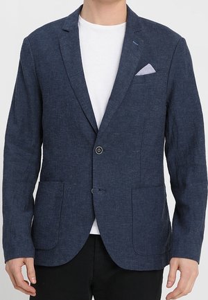 Mężczyzna ubrany w granatową marynarkę na jeden guzik z naszywanymi kieszeniami i jasnoniebieską poszetką, na białym t-shircie.