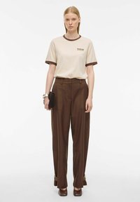 T-shirt beige a maniche corte con bordi marroni e logo; pantaloni marroni a vita alta con tasche laterali; la modella tiene una piccola pochette nera.