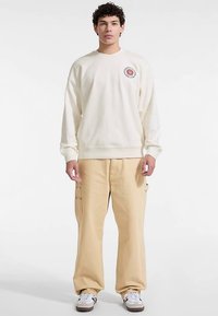 Sweat-shirt crème avec impression de logo, pantalon cargo beige coupe décontractée, et baskets blanches. Le tissu est doux, avec des poignets et une ceinture côtelés.
