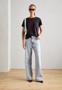 Donker t-shirt, lichte spijkerbroek met wijde pijpen, en puntige hakken. Accessoires omvatten een lichtblauwe tas en donkere zonnebril. Effen achtergrond.